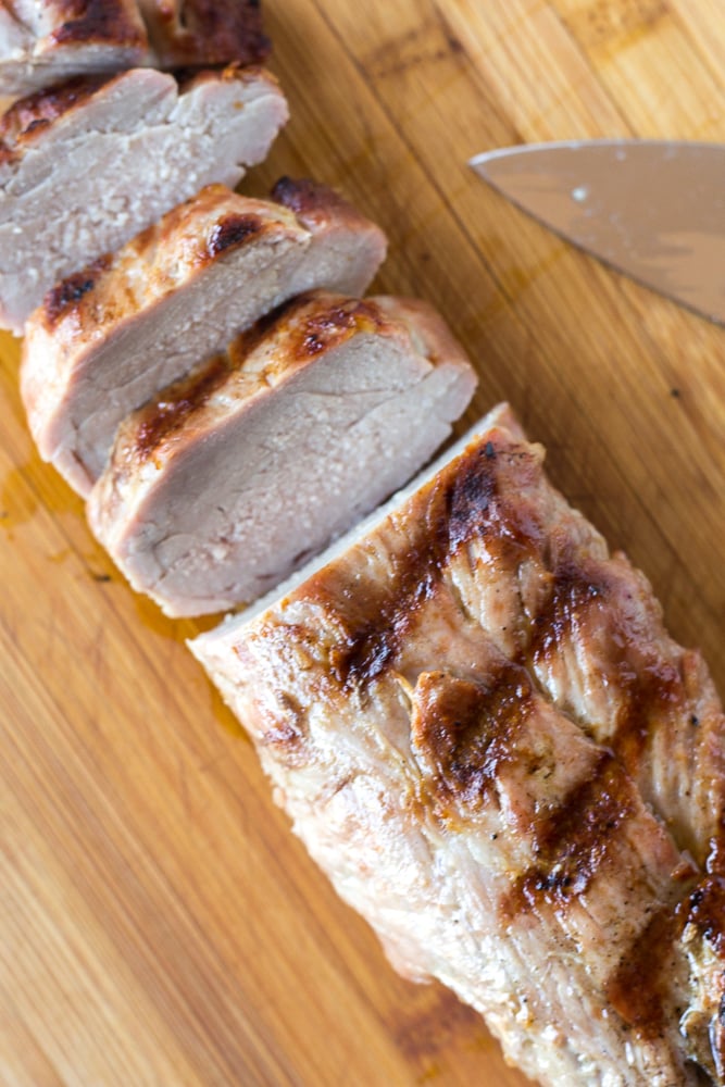 Grilled pork tenderloin being sliced on a wood cutting board.