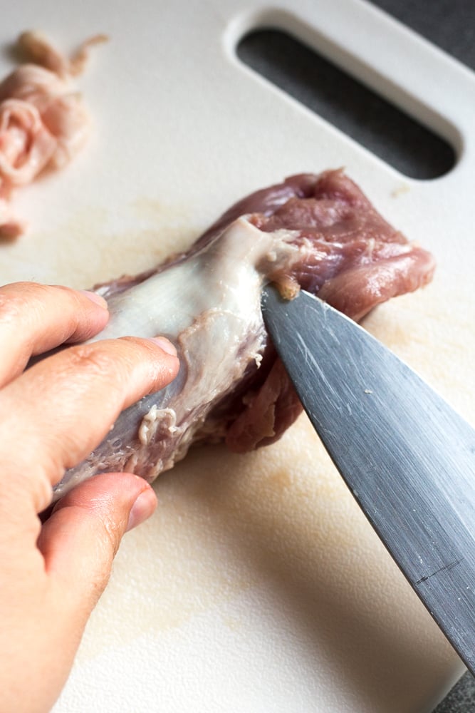 A large sharp knife cutting the silverskin off pork tenderloin on a white cutting board.