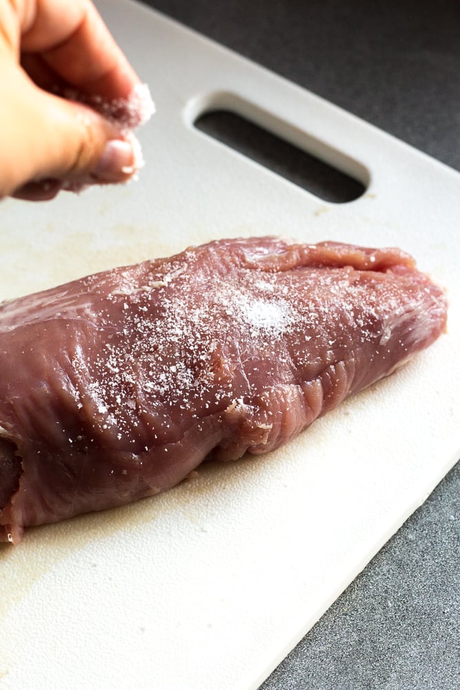 A hand sprinkling kosher salt on raw pork tenderloin that's on a white cutting board.