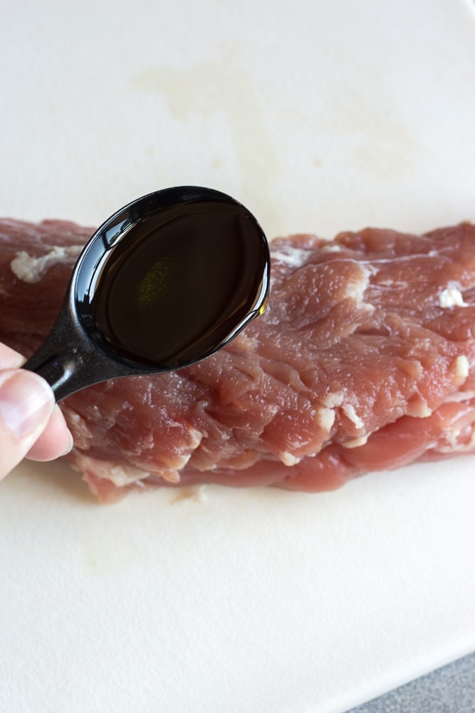 A hand pouring a tablespoon of oil on raw pork tenderloin resting on a white cutting board.