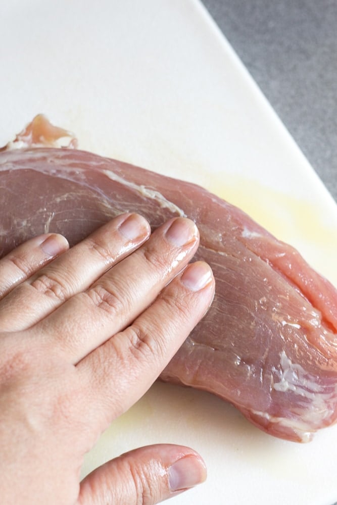 A hand rubbing oil onto raw pork tenderloin on a white cutting board.