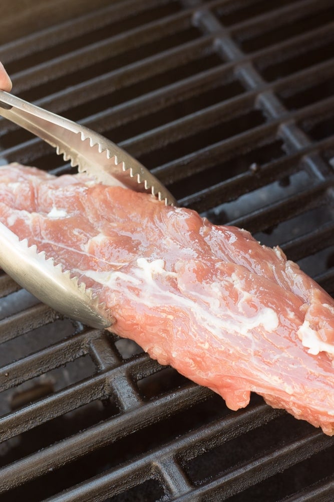 A pair of metal tongs placing brined pork tenderloin on the grill.