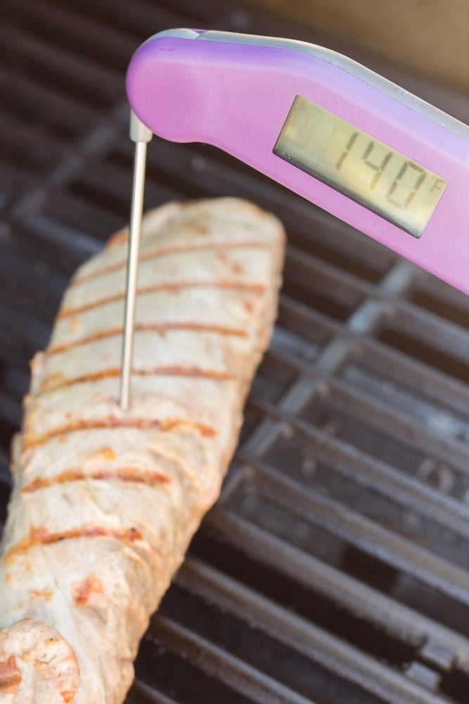 A purple meat thermometer checking the internal temperature of grilled pork on grill grates.