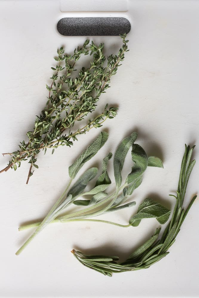 Fresh sage, rosemary, and thyme on a white cutting board.
