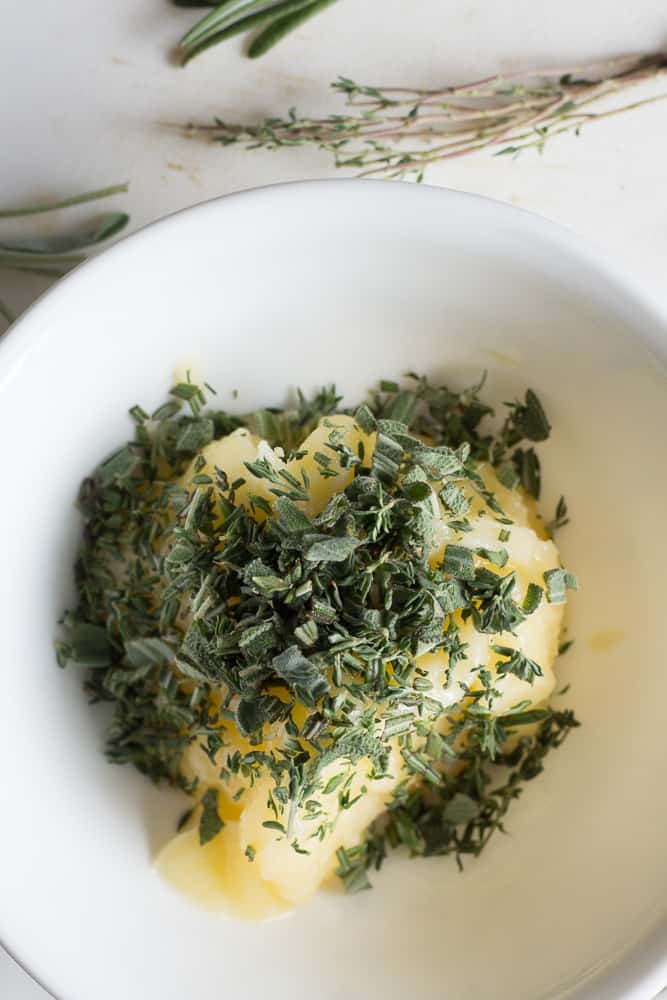 Chopped fresh herbs on top of softened ghee in a white bowl.