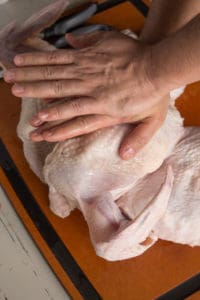 two hands pushing down on a spatchock turkey to help it flatten on a cutting board