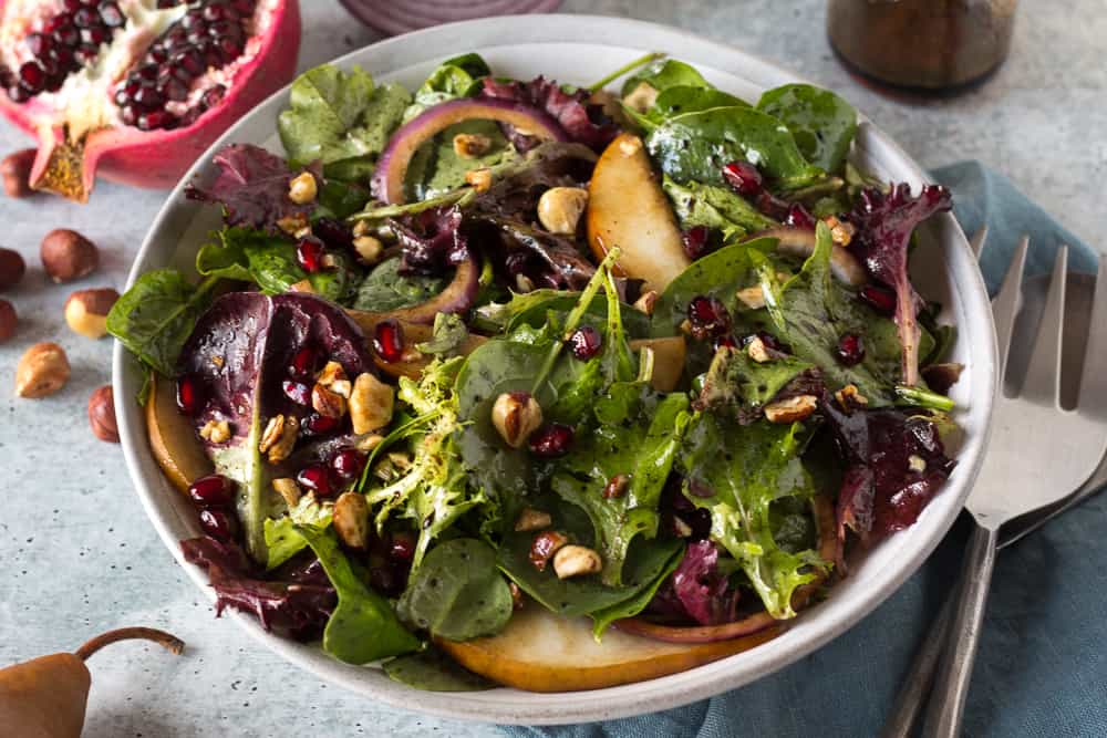 A close up of a green salad with pears, hazelnuts, red onion, and pomegranate in a gray bowl.