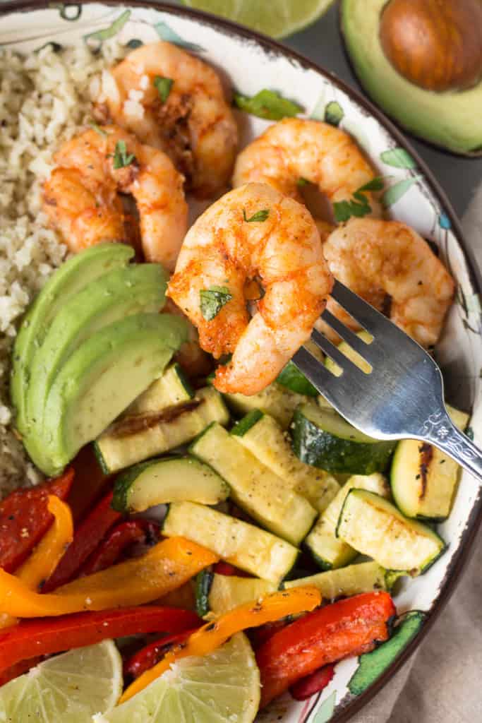 a fork holding a piece of grilled shrimp over a bowl of grilled veggies and shrimp