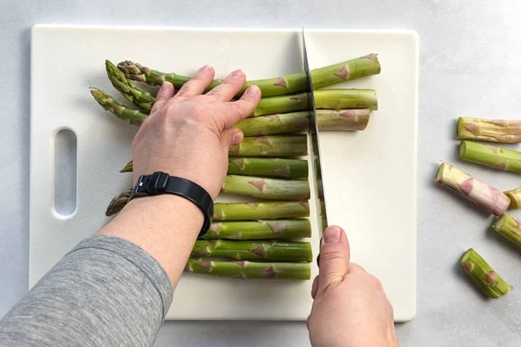 Two hands using a large sharp knife to cut the ends off a bunch asparagus stalks on a white cutting board.