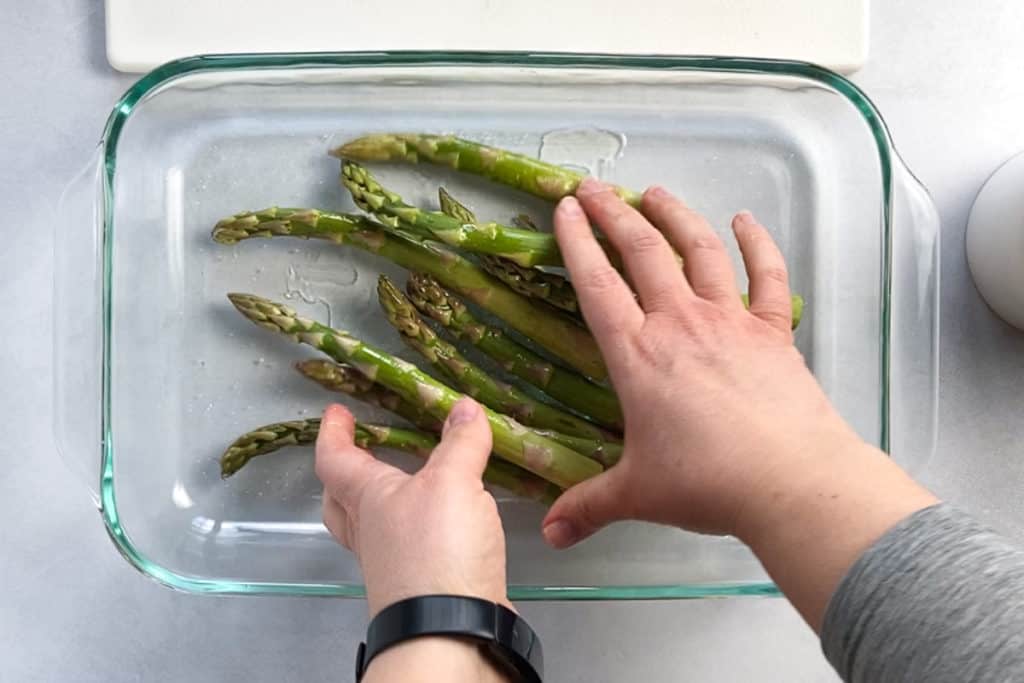 Two hands rubbing oil and kosher salt over fresh asparagus in a glass casserole dish.