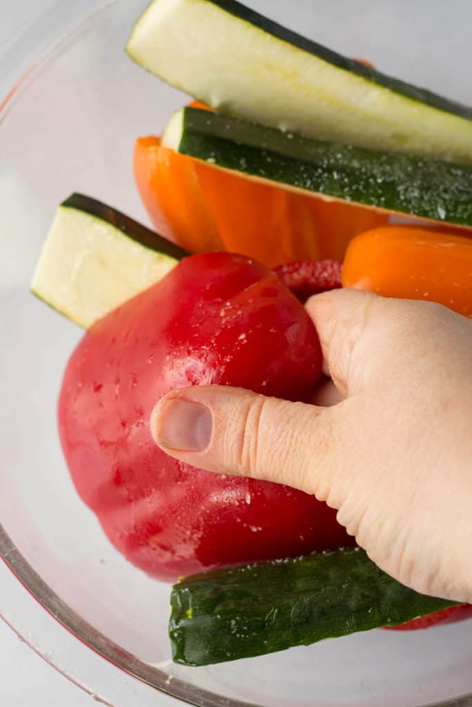 a hand rubbing oil on a bell pepper in a bowl with other veggies