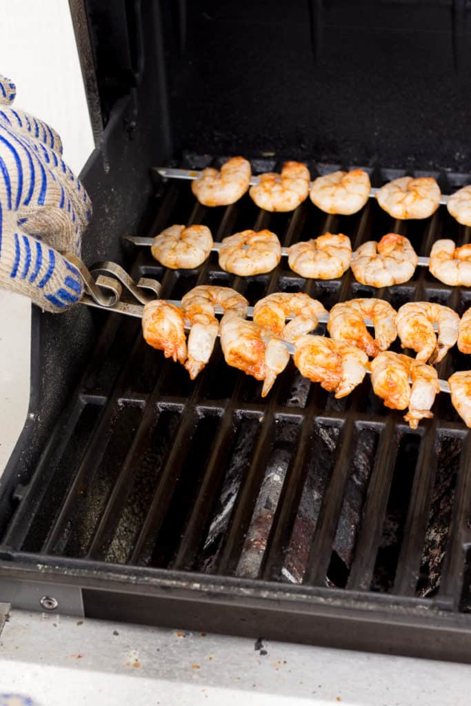 placing shrimp skewers on a gas grill with a hand in an oven mitt
