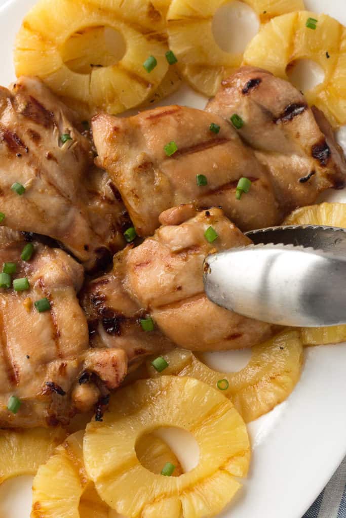 top down shot of grilled pineapple chicken on a white platter with tongs taking a piece