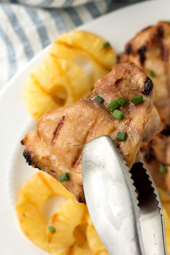 Close up of a pair of metal tongs holding a grilled chicken thigh above a white platter with chicken thighs and pineapple on it.