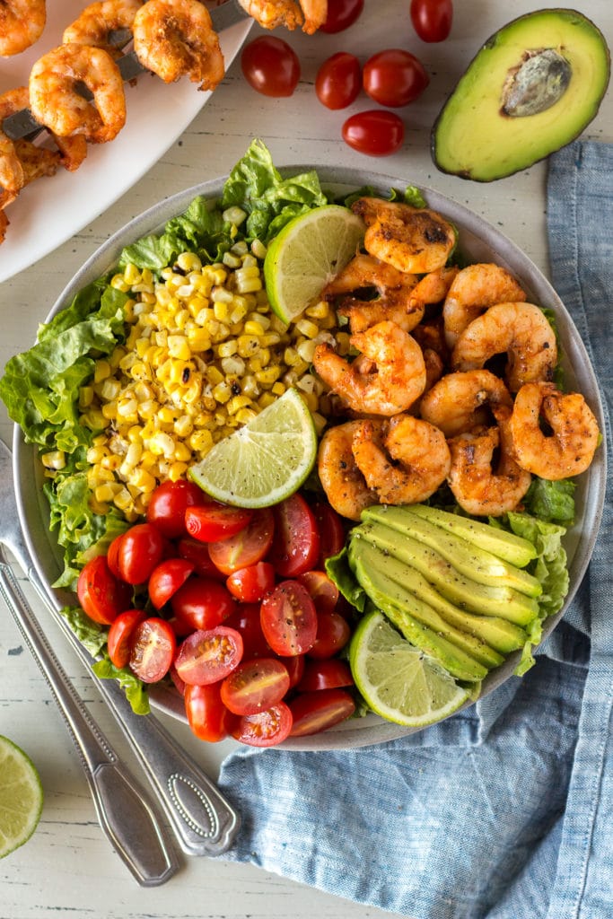 Top down shot of grilled shrimp and corn salad in a bowl with tomatoes, avocado, and slices of lime.
