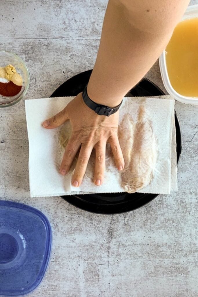 Top down shot of a hand drying off brined raw chicken breasts on a black plate with paper towels.