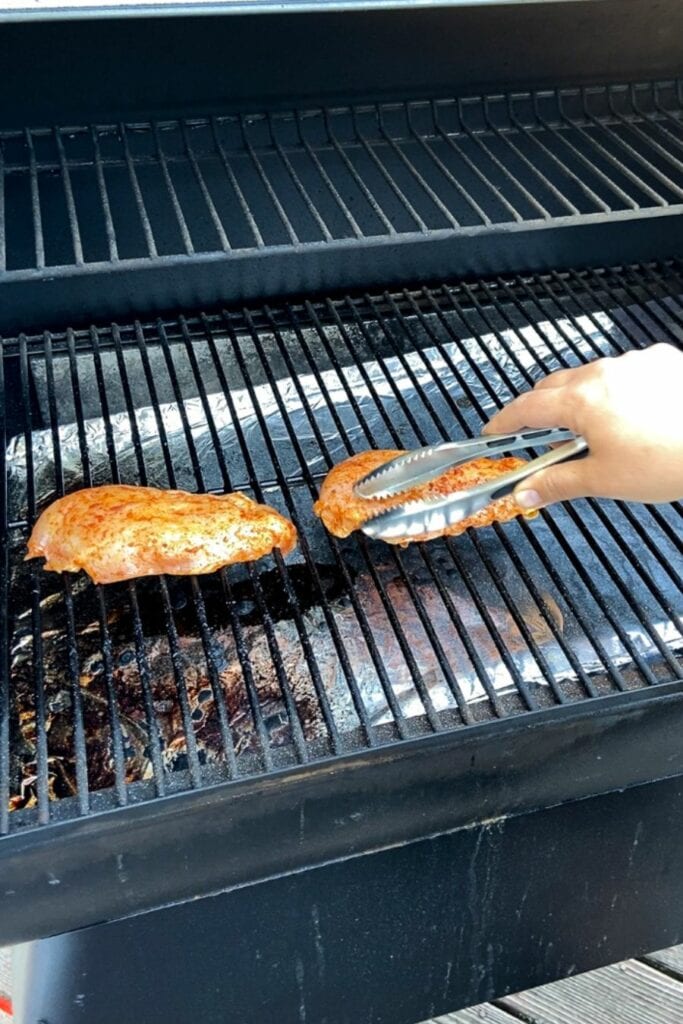 Tongs setting brined raw chicken breasts with seasoning on them onto grill grates.
