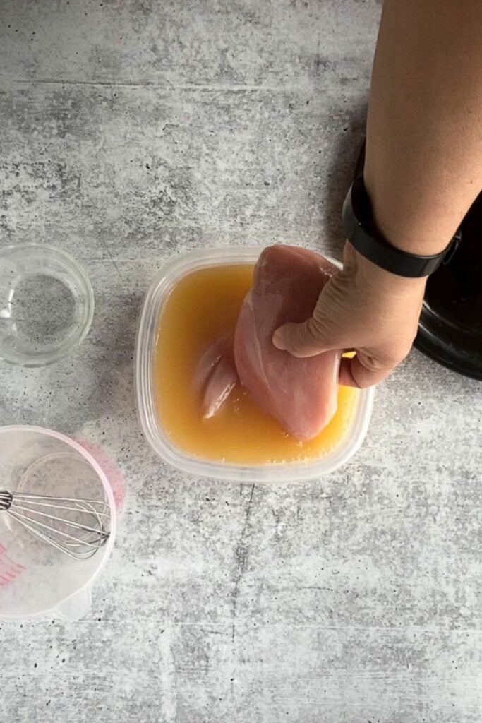 Top down shot of a hand placing a raw boneless chicken breast in a square plastic container with apple juice brine in it.
