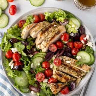 top down shot of greek salad with grilled chicken with a striped blue and white napkin and veggies to the side.