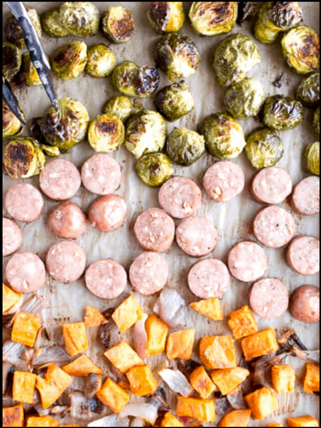 Top down view of a sheet pan with roasted sweet potatoes, red onions, sliced sausage, and Brussels sprouts on it.