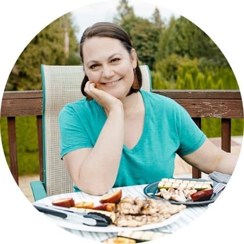 A woman with short brown hair wearing a teal t shirt sitting on a deck at a table with grilled food on plates in front of her.