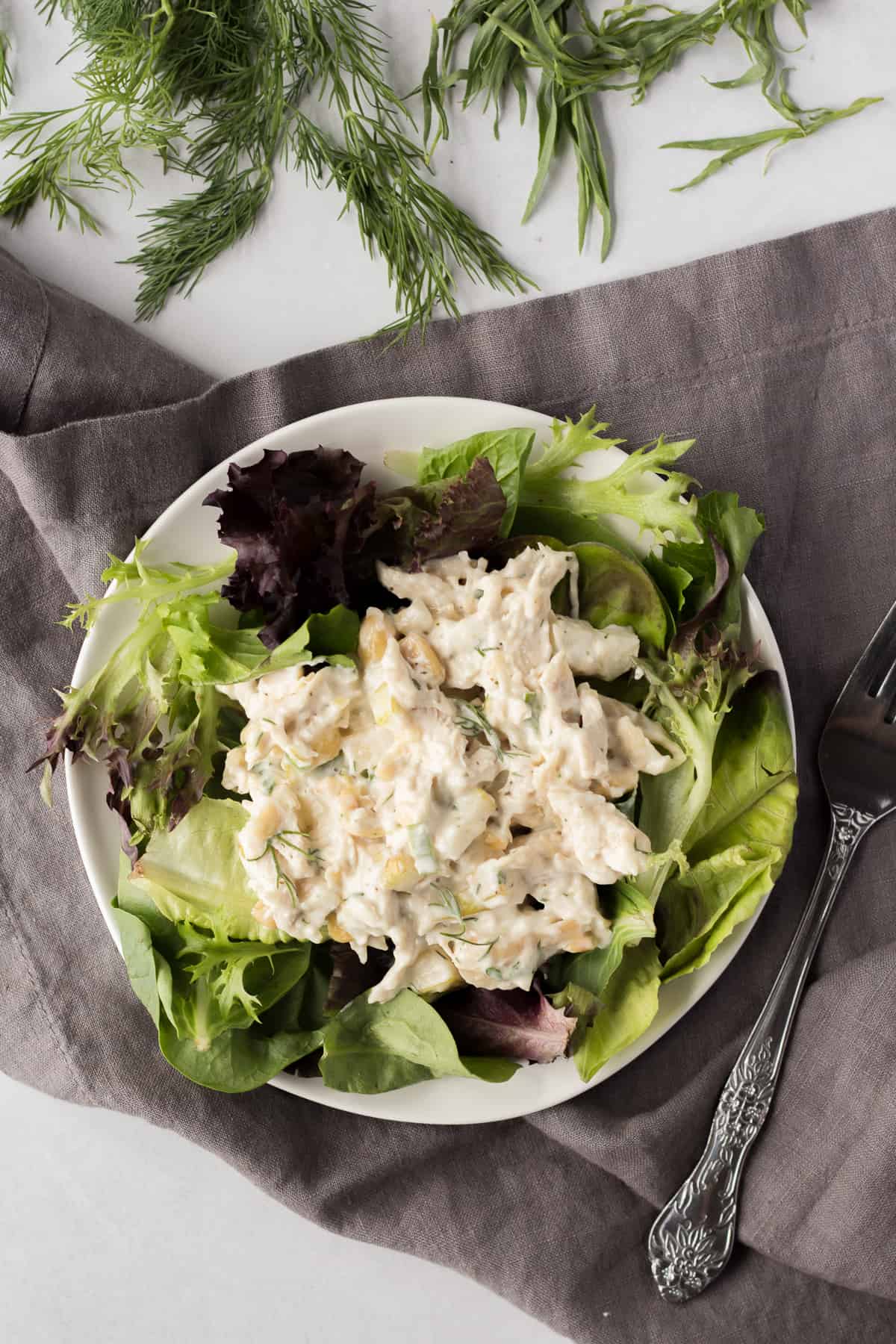 a white plate with a bed of greens and pear chicken salad on top over a gray napkin with herbs surrounding it