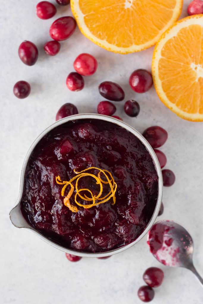 top down shot of gluten free cranberry sauce with an orange peel garnish in a small gray bowl. Fresh cranberries and slices of oranges surround it and a spoon dipped in cranberry sauce is on the bottom right.