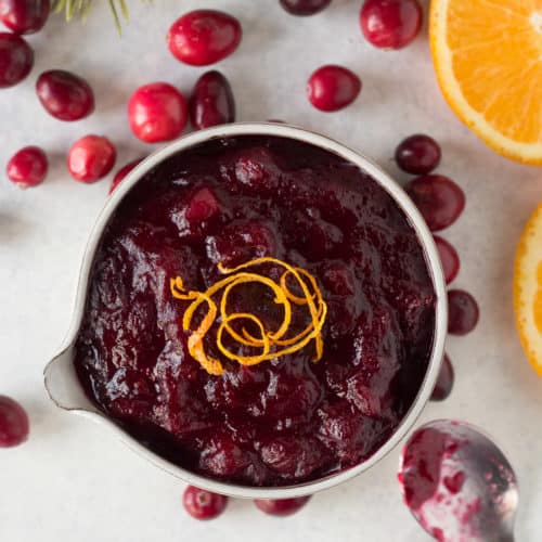 top down shot of cranberry sauce in a small gray bowl with orange zest on top, surrounded by fresh cranberries and some orange slices.