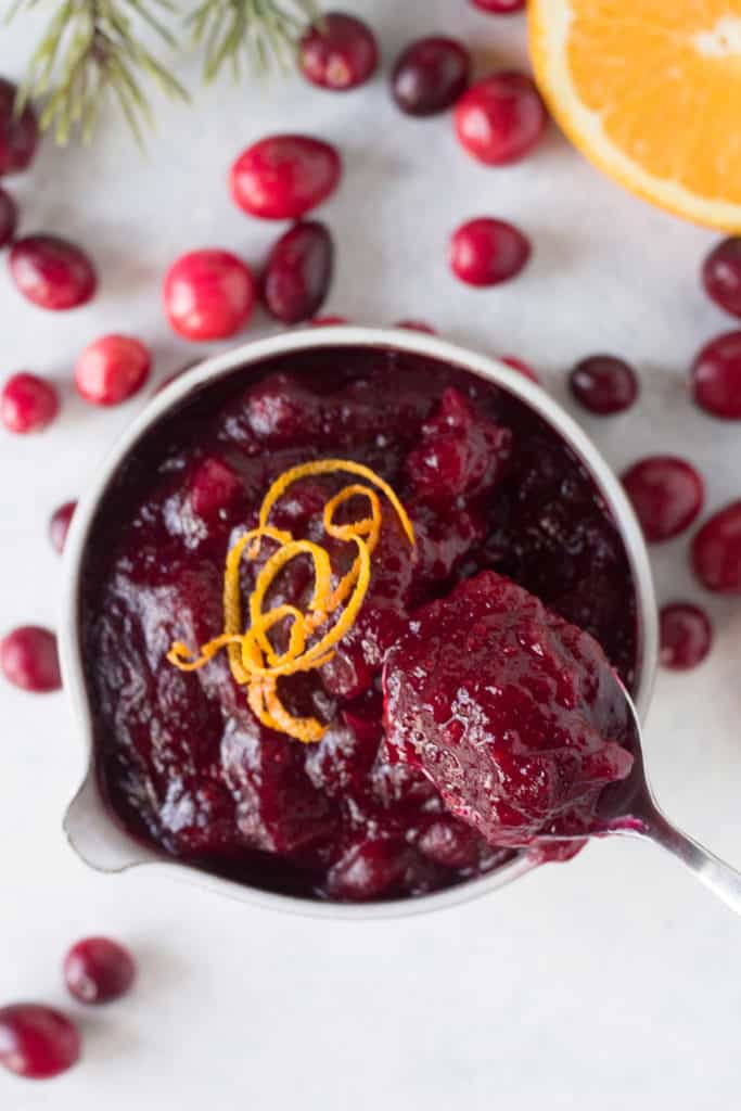 top down shot of a spoon being dipped into a small gray bowl filled with gluten free cranberry sauce. Fresh cranberries, a slice of orange, and some pine branches lay around the bowl.
