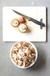 top down shot of a white cutting board with 3 mushrooms on it next to a bowl of sliced mushrooms.