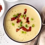 top down shot of a bowl of gluten free potato soup next to a tan napkin, a silver spoon, and two small white plates, one with green onion and the other with cooked bacon bits.