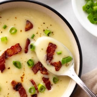 top down shot of a bowl of potato soup topped with bacon and green onions with a silver spoon dipping a bit out.