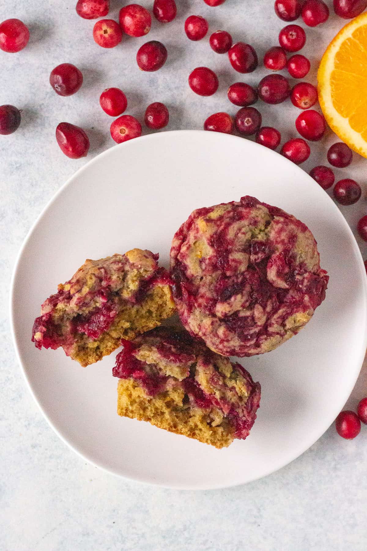 top down shot of two cranberry orange muffins on a white plate, one of which is cut in half. Fresh cranberries and an orange slice lie next to the plate.