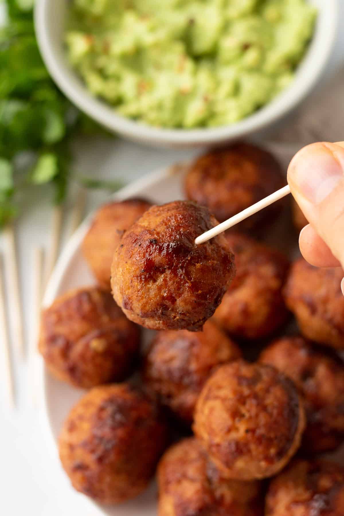 a hand holding a toothpick with a bbq turkey meatball on it. A plate of meatballs are in the background, with other toothpicks, greens, and a bowl of guacamole.