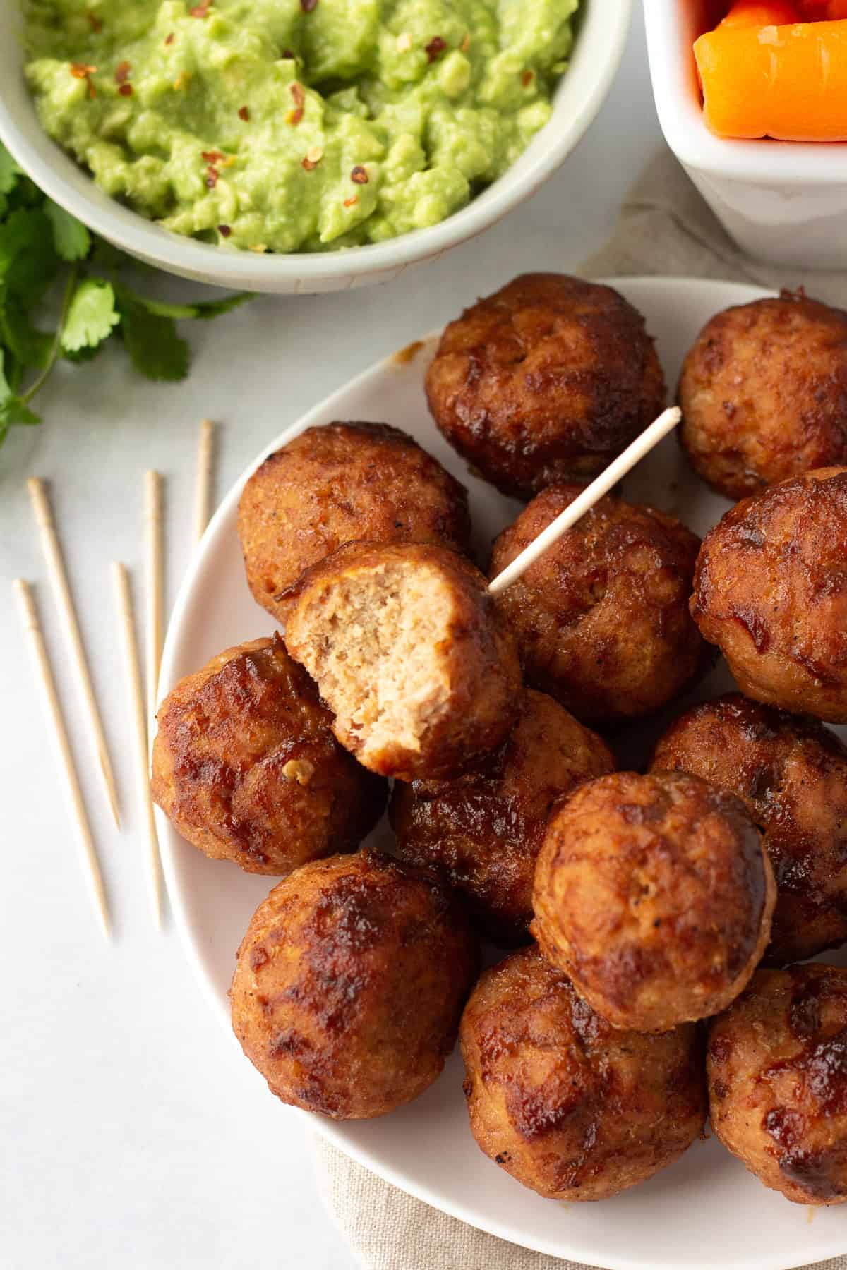 A white plate with bbq turkey meatballs piled on it. One has a toothpick in it and a bit taken out of it. Other toothpicks, guacamole, and carrots surround the plate of meatballs.