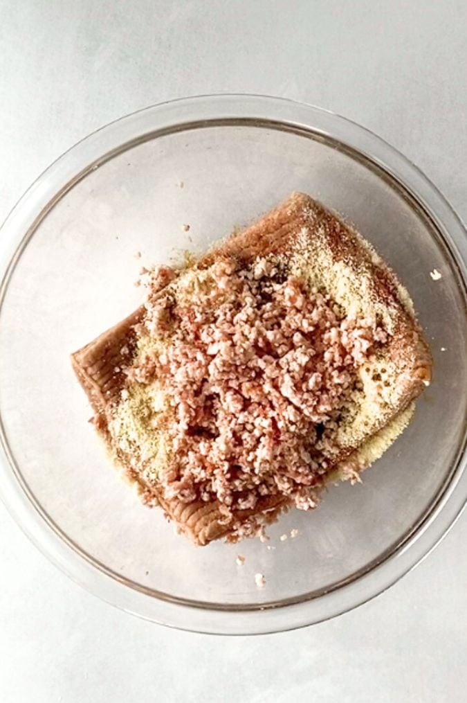 Top down shot of a pound of ground turkey with almond flour, minced bacon, and spices on it in a large clear bowl, all on top of a white surface.