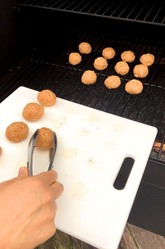 A pair of tongs placing raw turkey meatballs from a white cutting board onto a Traeger grill.