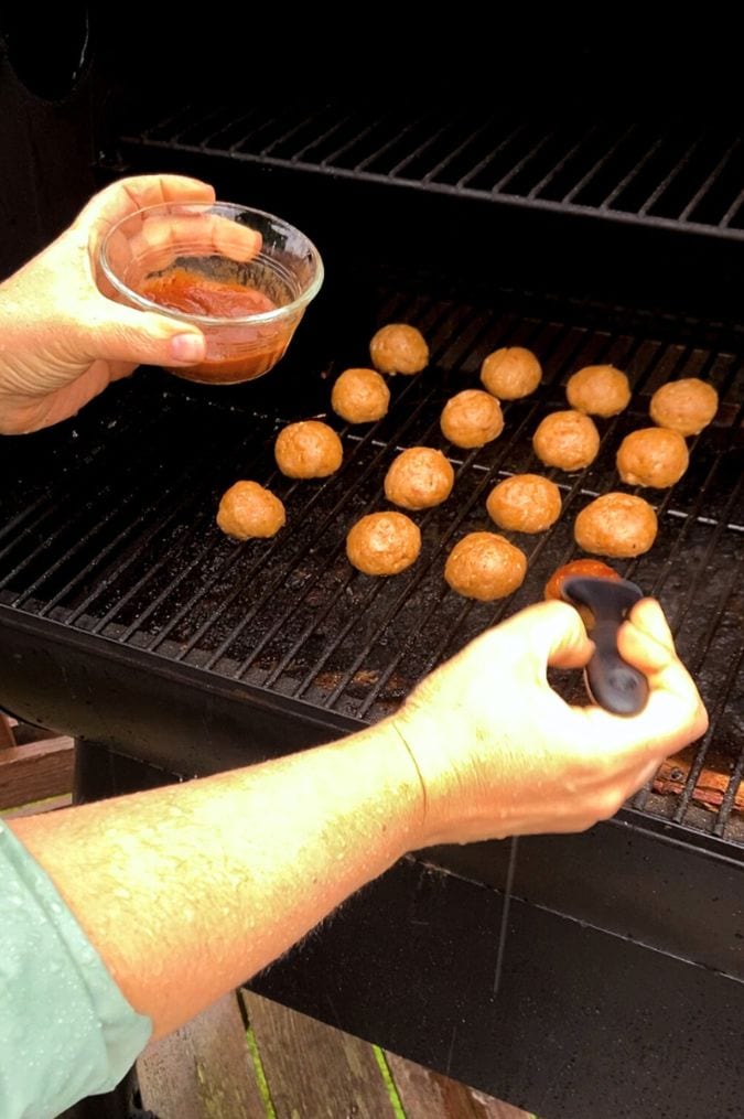 Hands brushing BBQ sauce onto smoked turkey meatballs on a grill.