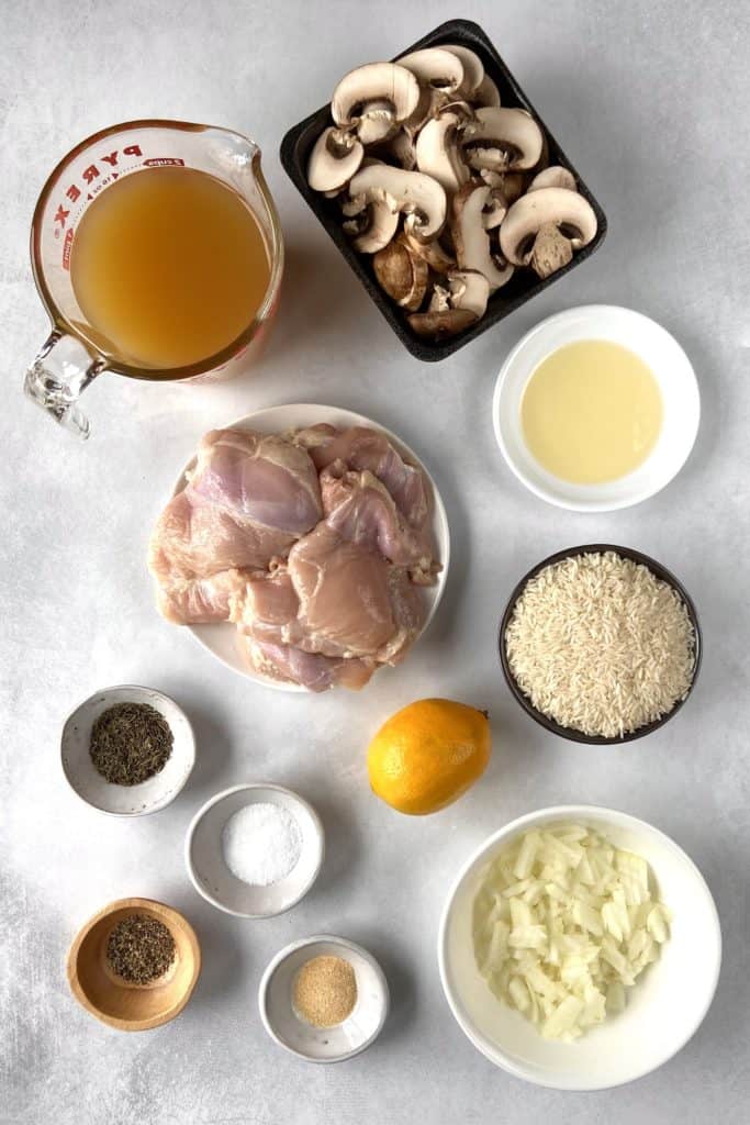 top down shot of ingredients for instant pot chicken thighs and rice, including sliced mushrooms, broth, oil, white rice, raw boneless skinless chicken thighs, a lemon, diced onion, and spices.
