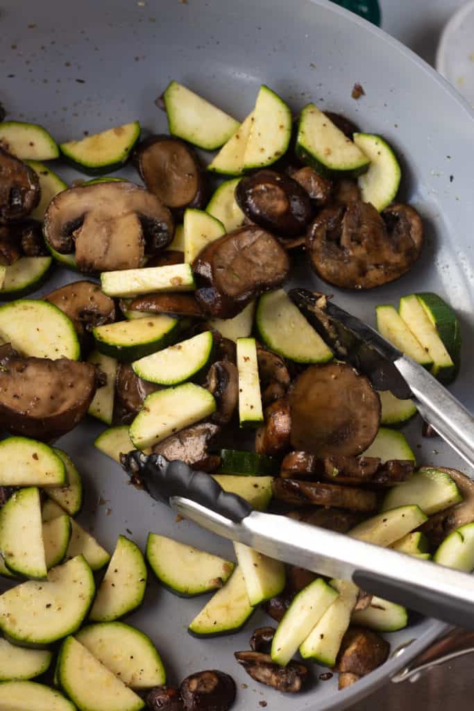 sauteed mushrooms and zucchini in a gray pan being stirred with tongs.