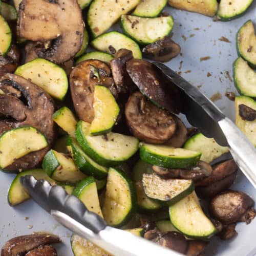 Zucchini and mushrooms in a gray pan being stirred with black tongs.