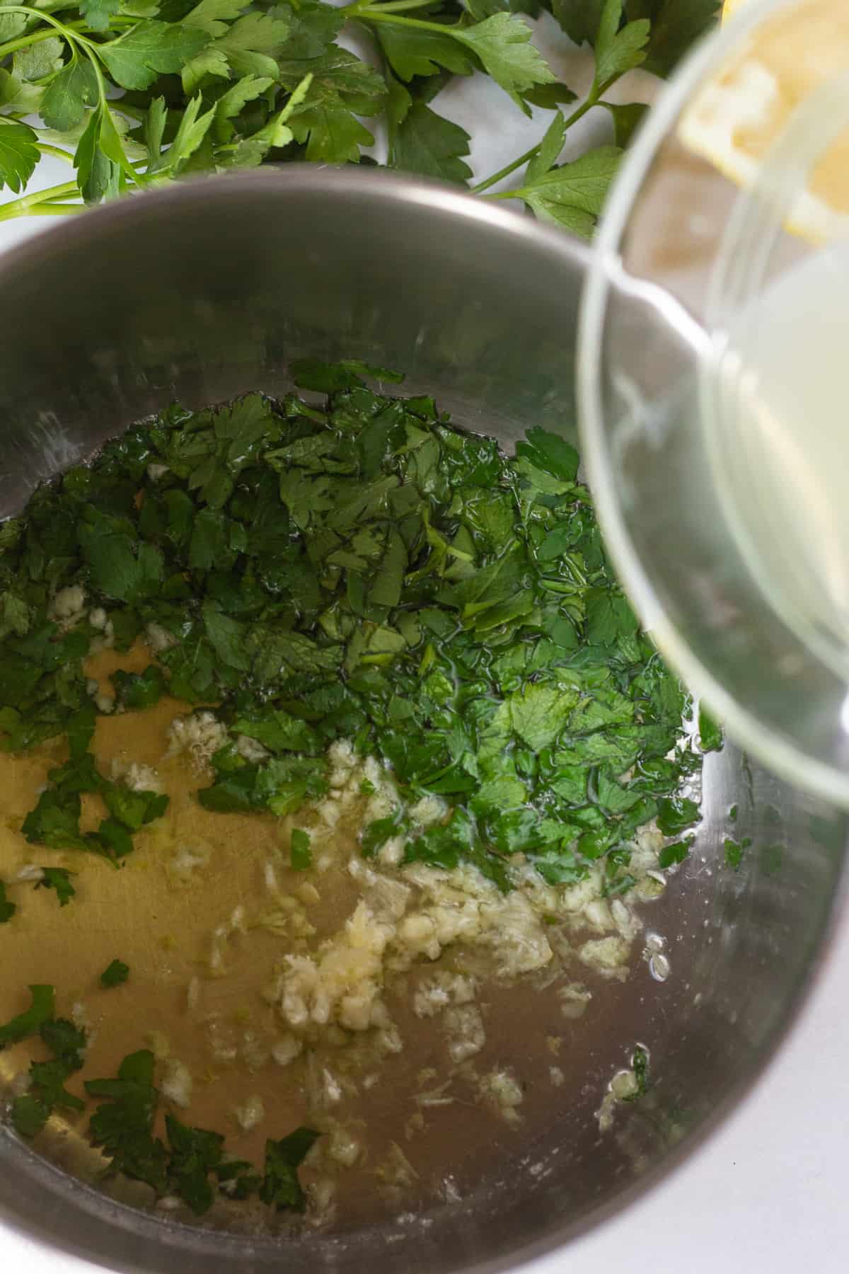 Top down shot of a small saucepan with ghee, chopped parsley, and minced garlic in it. Lemon juice is about to be poured from above out of a clear small dish.