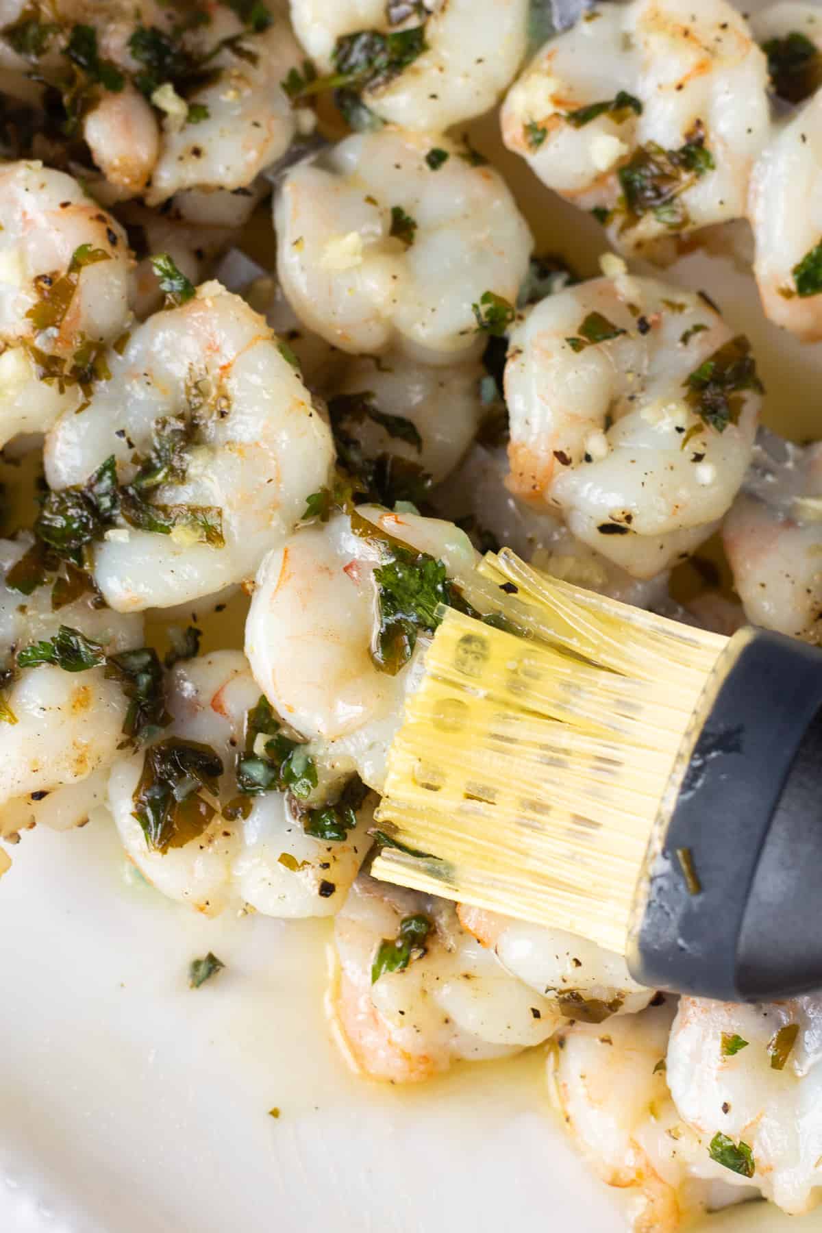 Garlic sauce being brushed onto shrimp skewers on a white platter by a silicone brush.