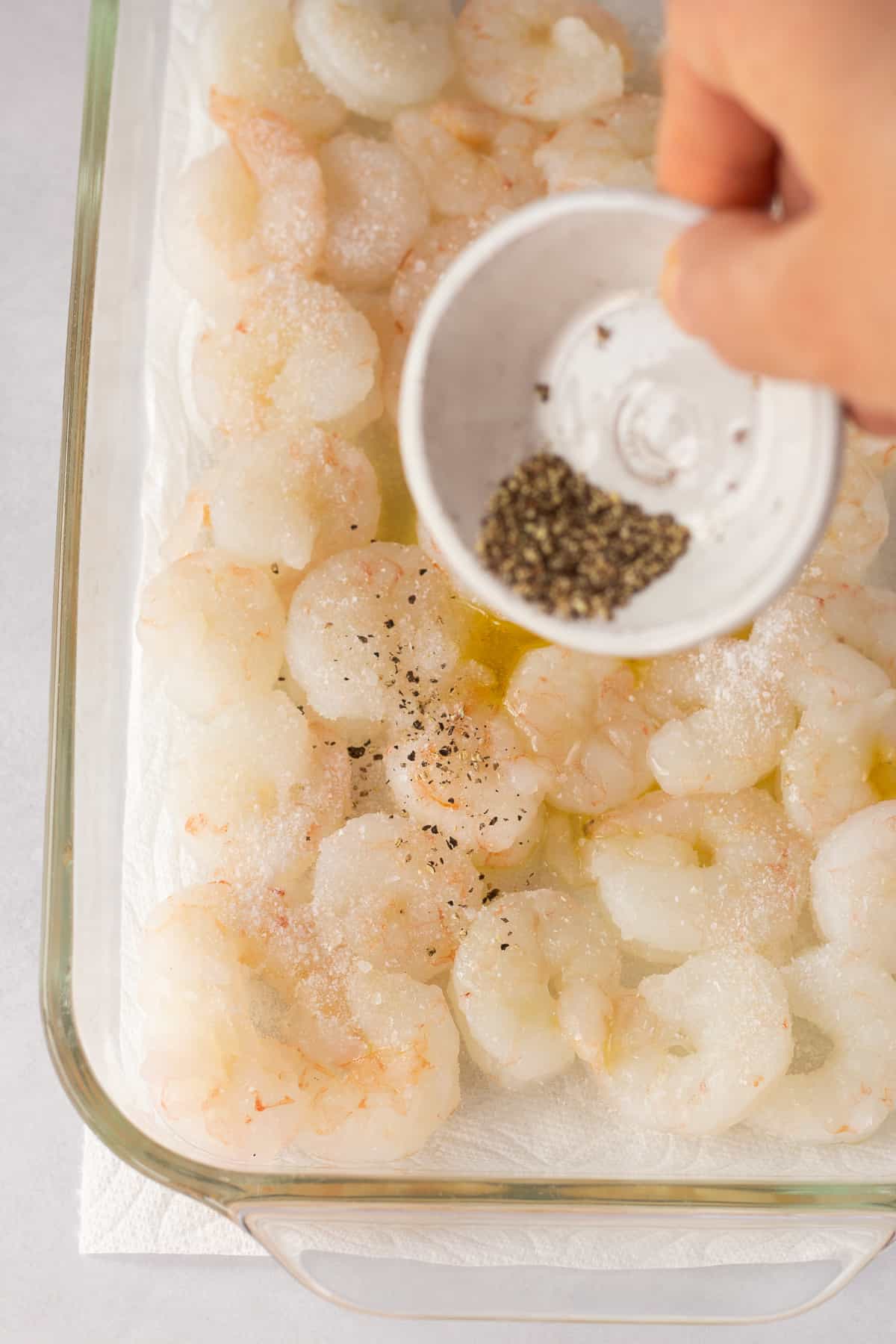 Pouring ground black pepper from a small white bowl into a clear dish with raw shrimp.