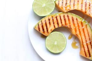 Close up of grilled cantaloupe drizzled with honey with cut limes on the side, all on a white plate.