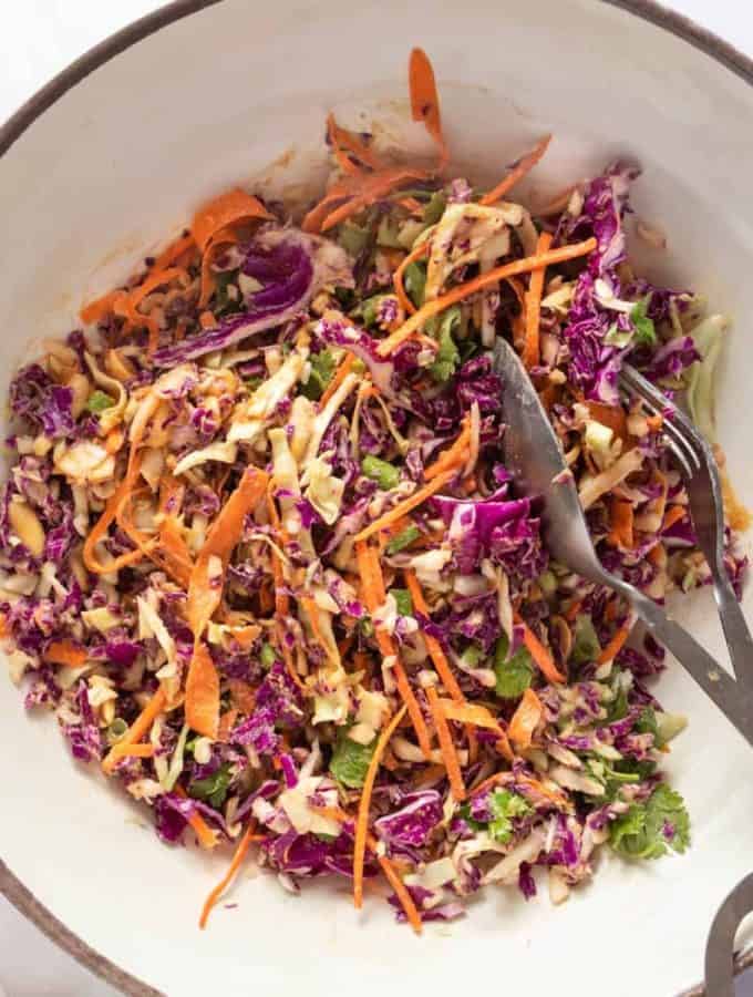 A slaw with purple and green cabbage and carrots with a nutty dressing in a large white bowl with a serving utensil in it.