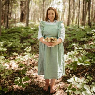 A woman standing in a forest wearing a green dress and holding a tray of food in her hands.