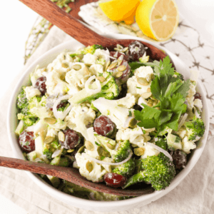 A raw broccoli and cauliflower salad with seeds, grapes, and fresh herbs in a white bowl.