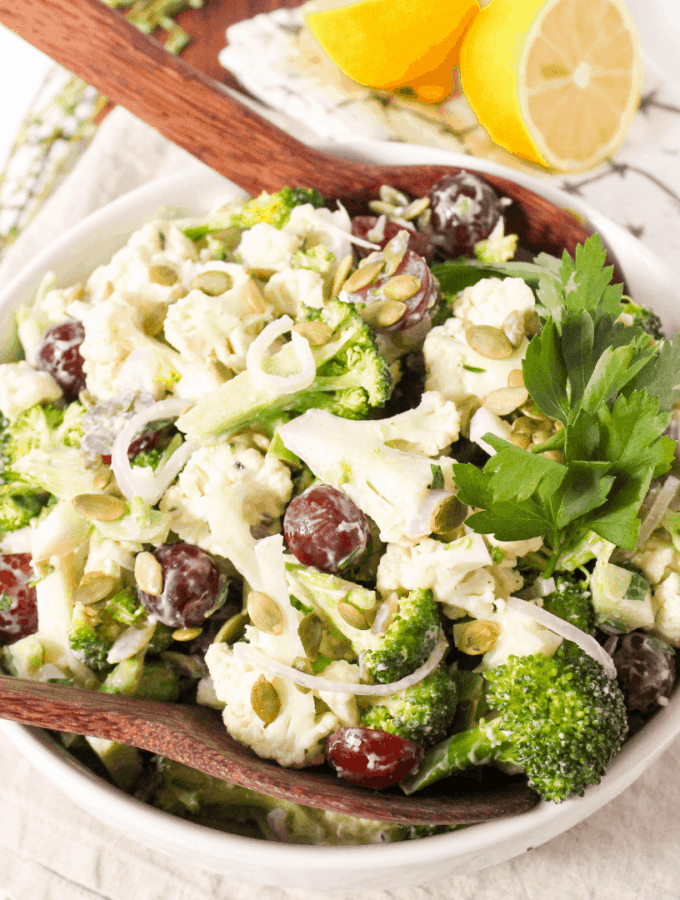 A raw broccoli and cauliflower salad with seeds, grapes, and fresh herbs in a white bowl.