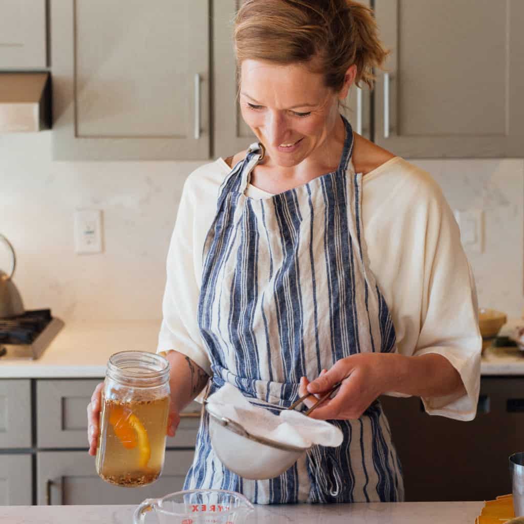 A woman with blonde hair in a kitchen, wearing a white shirt and blue and white striped apron. She's holding a mason jar with liquid in one hand and a strainer with cheesecloth in the other.